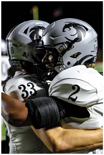 Two people wearing football helmets and uniforms are embracing on a field during a game. Description generated by AI