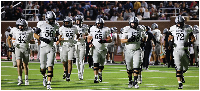 Six football players in white and gray uniforms with helmets walk together on a field with a crowd in the background. Description generated by AI
