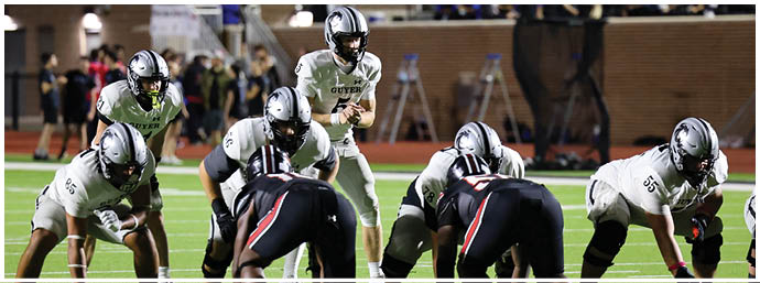 A group of people in football uniforms are positioned on a field preparing for a play during a game with spectators in the background. Description generated by AI
