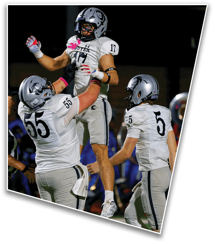 Three football players in white uniforms and helmets celebrate on the field with one player being lifted by two others. Description generated by AI