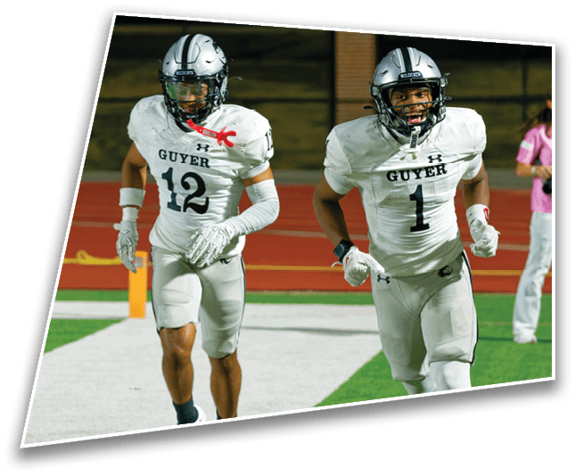 Two people in white football uniforms with helmets are running on a field while a person in casual clothing stands in the background near the track. Description generated by AI
