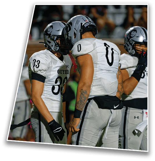 Two football players in white uniforms with helmets are standing close together while another player in a black uniform stands nearby on a field. Description generated by AI