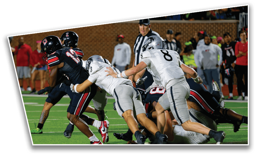 Several people in football uniforms are engaged in a play on a field with a referee and spectators in the background. Description generated by AI