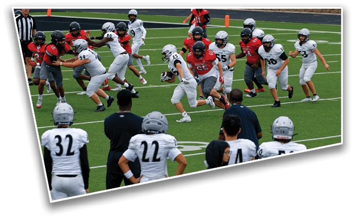 A group of people wearing football helmets and uniforms are actively playing on a green field while others watch from the sidelines. Description generated by AI