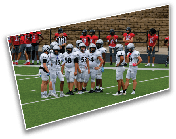 A group of people in white football uniforms stand together on a field while another group in red uniforms stands in the background near a brick wall. Description generated by AI