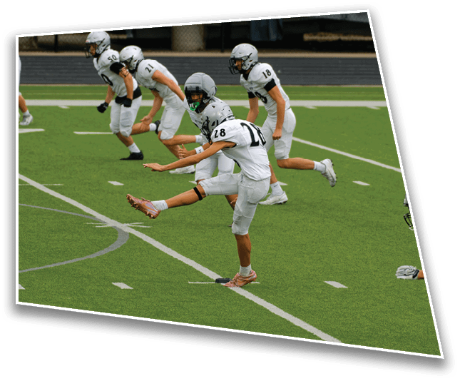 A person in a white football uniform is kicking a football while several other players in similar uniforms stand behind on a green field with white yard lines. Description generated by AI