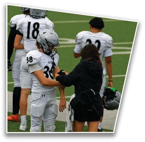 A person in a black jacket is adjusting the arm of a person wearing a white football uniform and helmet on a sports field with two other players in white uniforms standing nearby. Description generated by AI