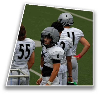 Four people wearing white football uniforms and helmets stand on a green field, with one person facing forward and the others facing away. Description generated by AI