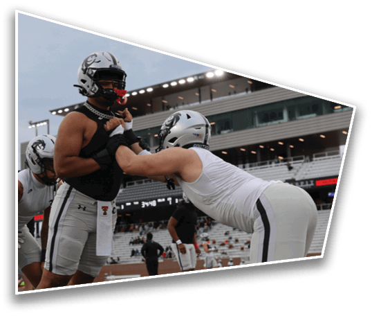 Two people wearing football helmets and athletic gear are engaged in a blocking drill on a field with a stadium in the background. Description generated by AI