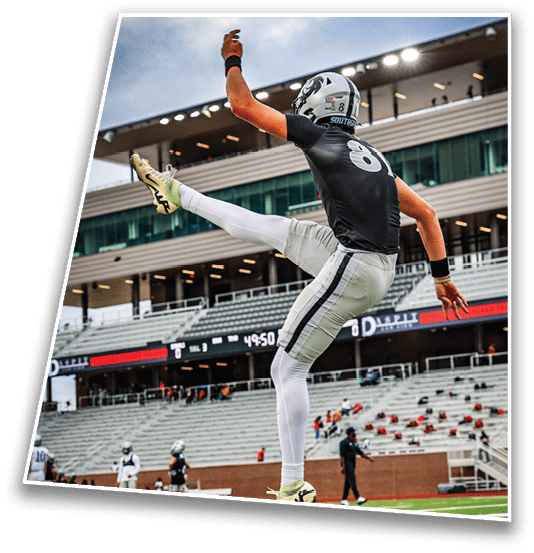 A person in football gear is jumping high on a football field with a stadium and other players in the background. Description generated by AI
