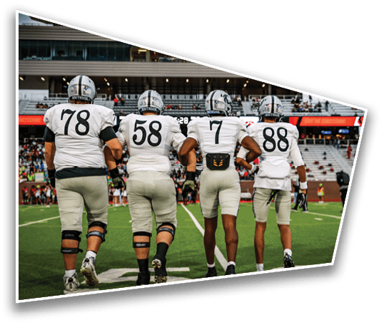 Four football players in white uniforms walk arm in arm on a field toward the stands during a game. Description generated by AI