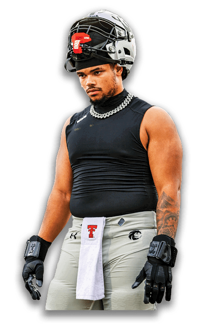 A person wearing football gear including a helmet, sleeveless black shirt, gloves, and light-colored pants with a towel tucked in the waistband stands with a focused expression. Description generated by AI