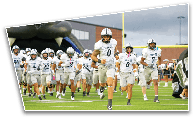 A group of adults in white football uniforms with helmets are running onto a green field from a large inflatable tunnel under a cloudy sky. Description generated by AI