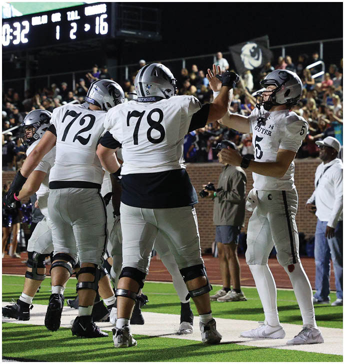 Three football players in white uniforms celebrate on the field near a scoreboard showing the game time and score with a crowd in the background. Description generated by AI