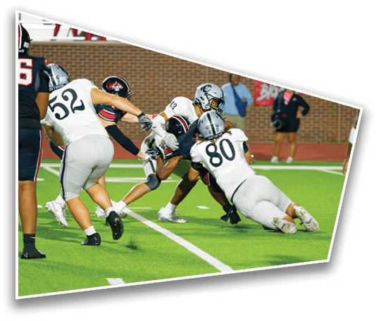 Several people in football uniforms are engaged in a tackle on a green field with a brick wall and spectators in the background. Description generated by AI