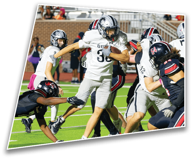 People in football uniforms are actively engaged in a game on a green field with one person holding the ball while others attempt to tackle or block. Description generated by AI