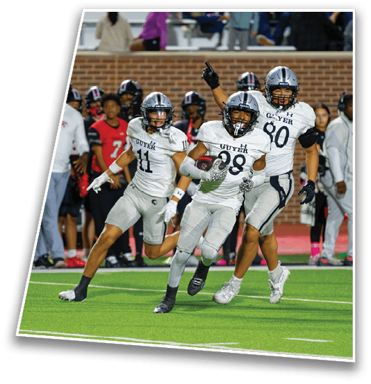 Three football players in white uniforms with helmets are running on a field with one player holding a football and another pointing upward while people in red and black uniforms stand in the background. Description generated by AI
