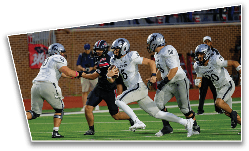Four football players in white uniforms are running on a field with one holding the ball while two players in black uniforms attempt to block them. Description generated by AI