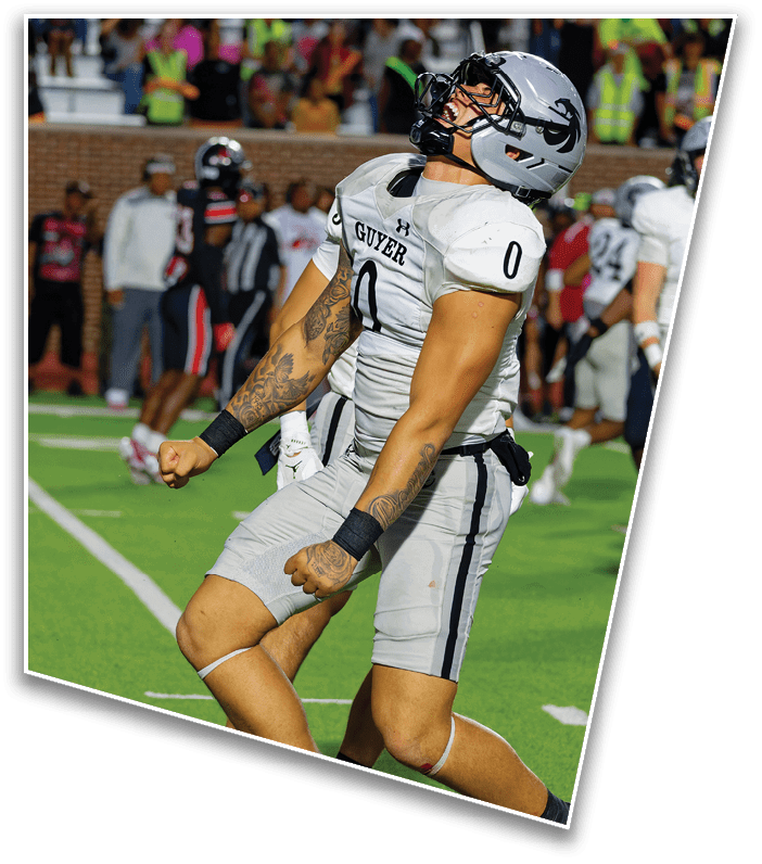 Two football players in gray uniforms with black stripes and helmets celebrate on a green field during a game. Description generated by AI