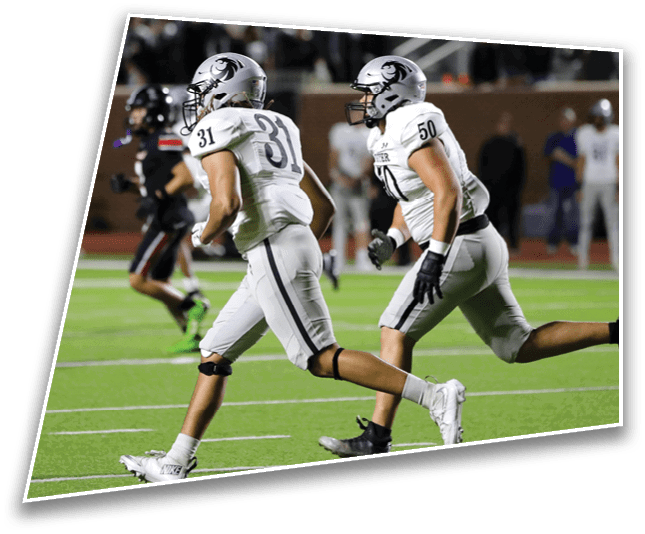 Two people in white football uniforms are running on a green field during a game. Description generated by AI