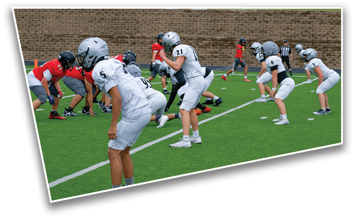 Five people in football uniforms and helmets stand on a field giving thumbs up, with others and buildings visible in the background. Description generated by AI