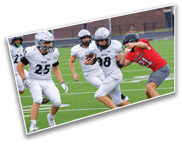 Four people in football uniforms are actively playing on a green field with one person in white running with the ball while another in red attempts to block. Description generated by AI
