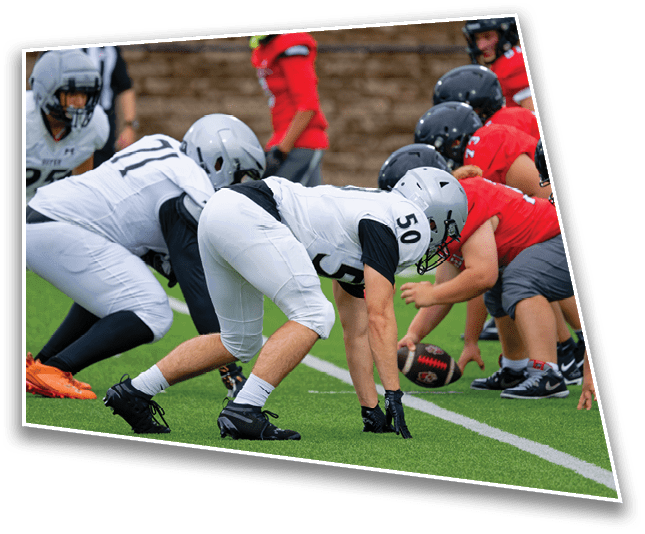 Two opposing football teams in helmets and uniforms are lined up facing each other on a green field, preparing for a play. Description generated by AI