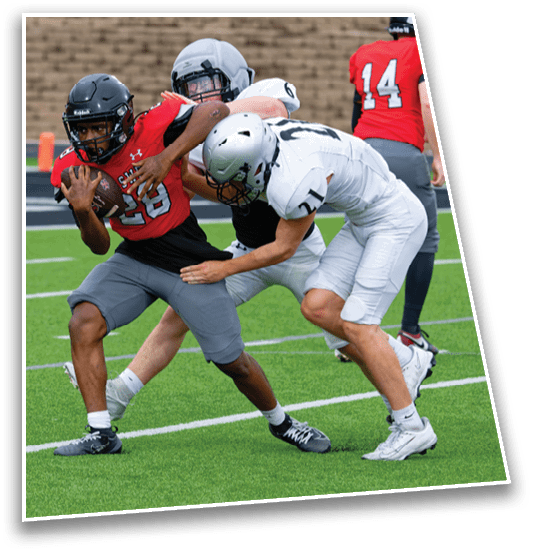 An adult in a red sports jersey is running with a football while two adults in white sports uniforms attempt to tackle them on a green football field. Description generated by AI