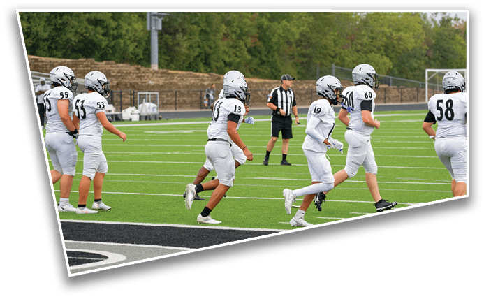 Several people in white football uniforms and helmets are running on a green football field near the end zone with a referee in the background. Description generated by AI