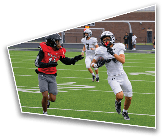 Two people in football uniforms are actively running on a field with one person holding a football while another reaches out toward them. Description generated by AI