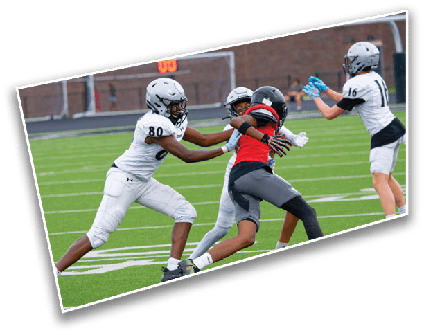 Two people in football uniforms are engaged in a play on a green field with a scoreboard and goalposts in the background. Description generated by AI