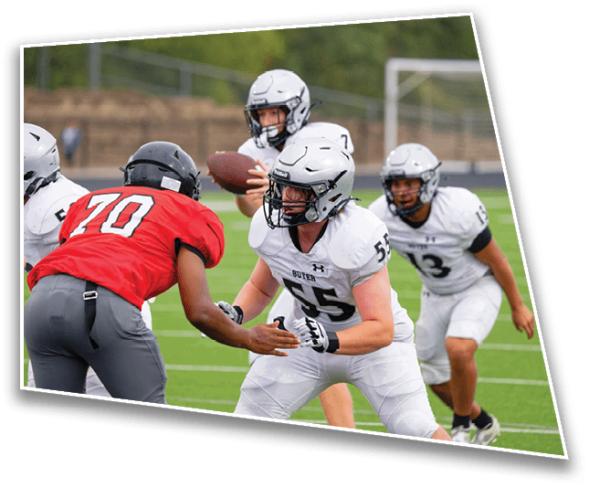 Four people in football uniforms are engaged in a play on a green field with one person holding a football and others preparing to block or defend. Description generated by AI