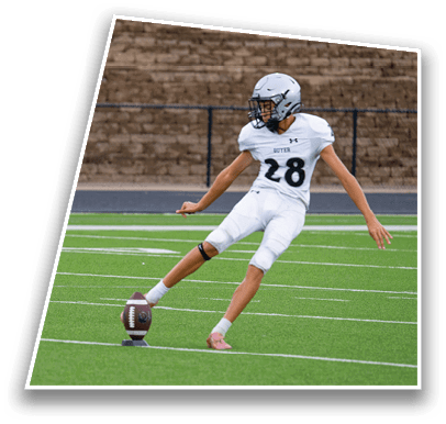 A person in a white football uniform is kicking a football on a green field with a stone wall in the background. Description generated by AI