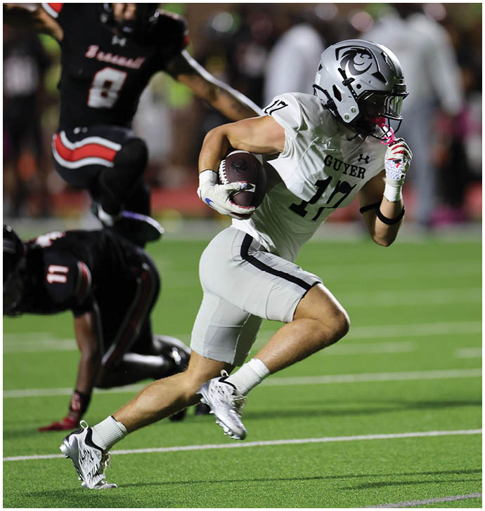 A person in a white football uniform with the word “GUYER" is running on a field while holding a football and being pursued by players in black uniforms. Description generated by AI