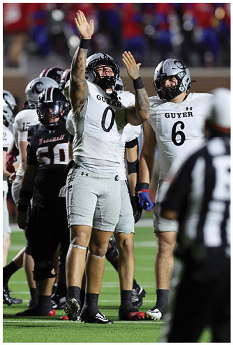 Two football players in white uniforms with helmets are celebrating on the field while other players and a referee stand nearby. Description generated by AI