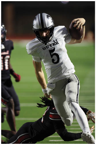 A person wearing a white football uniform with the number 5 is running with the ball while another person in a black uniform attempts to tackle them on a football field. Description generated by AI