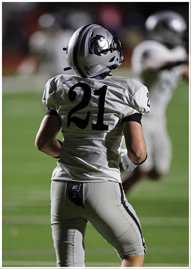 A person wearing a white football uniform with the number 21 on the back is standing on a green field during a game. Description generated by AI