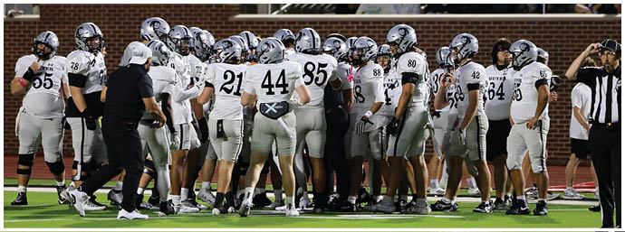 A group of people wearing white and black football uniforms are gathered on a field with a crowd seated in the background. Description generated by AI