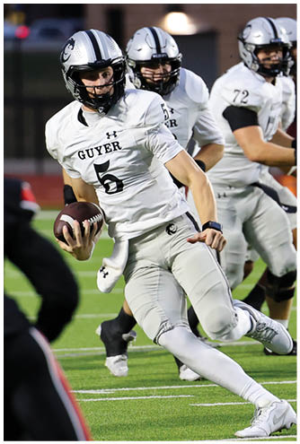 A person in a white football uniform with the number 5 is running on a field while holding a football, with two other players in similar uniforms in the background. Description generated by AI