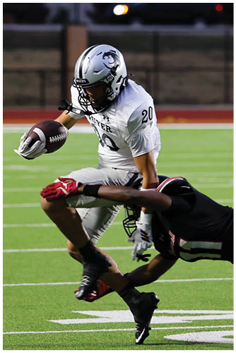 A person in a white football uniform is running with the ball while another person in a black football uniform is diving to tackle them on a green field. Description generated by AI