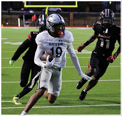A person in white sports gear is running with a football while another person in black sports gear attempts to catch them on a football field with a referee in the background. Description generated by AI