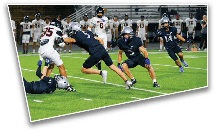 People in football uniforms are actively playing on a field with some players tackling and others running, while additional players stand in the background. Description generated by AI