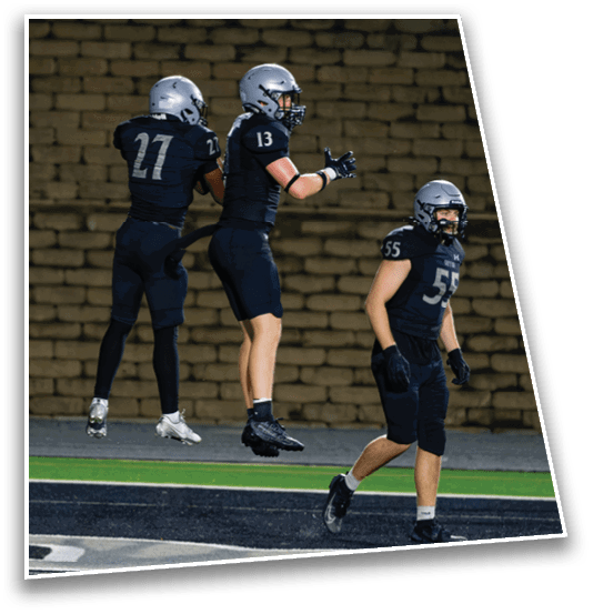 Three people in football uniforms and helmets are on a field with two of them jumping in the air near a brick wall background. Description generated by AI