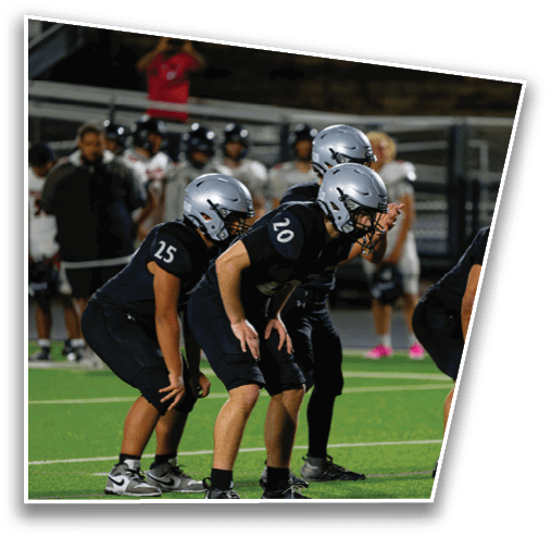 Five people wearing black football uniforms and helmets are crouched in a ready position on a green field with other players and a coach visible in the background. Description generated by AI