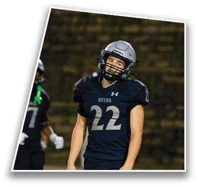 A person wearing a dark football uniform with the number 22 and the word Guyer on the chest stands on a field with another player partially visible in the background. Description generated by AI