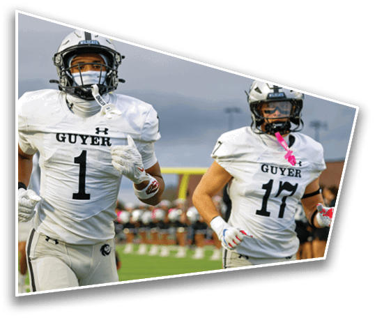 Two people in white football uniforms with helmets are running on a field during a game, with one wearing a jersey labeled “GUYER 17." Description generated by AI