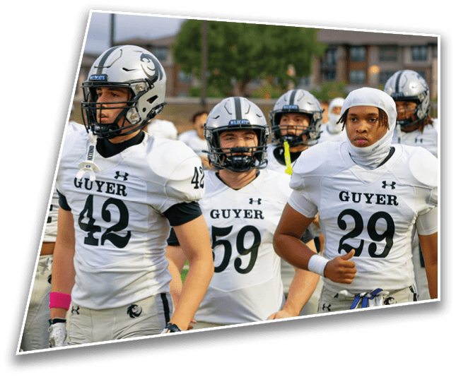 A group of people wearing white football uniforms with helmets and numbers stand together on a field with a blurred background of trees and buildings. Description generated by AI