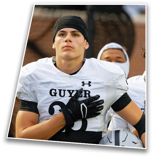 A person wearing a white football jersey with the word “GUYER" and the number 26 stands with one hand over their chest and a black head covering. Description generated by AI