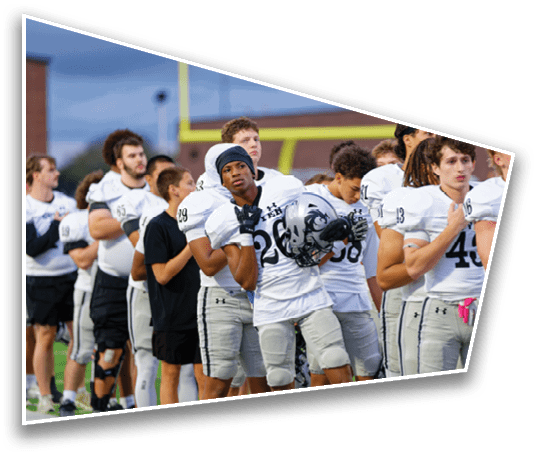 A group of people wearing white football uniforms stand closely together on a field, some holding helmets and others with hands over their chests. Description generated by AI