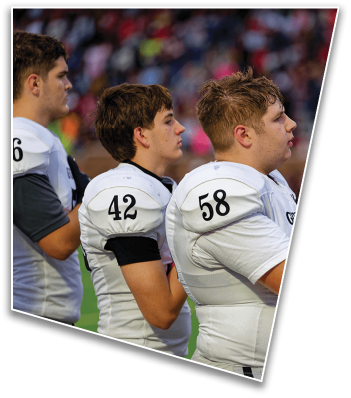 Two people in white football uniforms stand side by side on a field with one person placing a gloved hand over their chest. Description generated by AI
